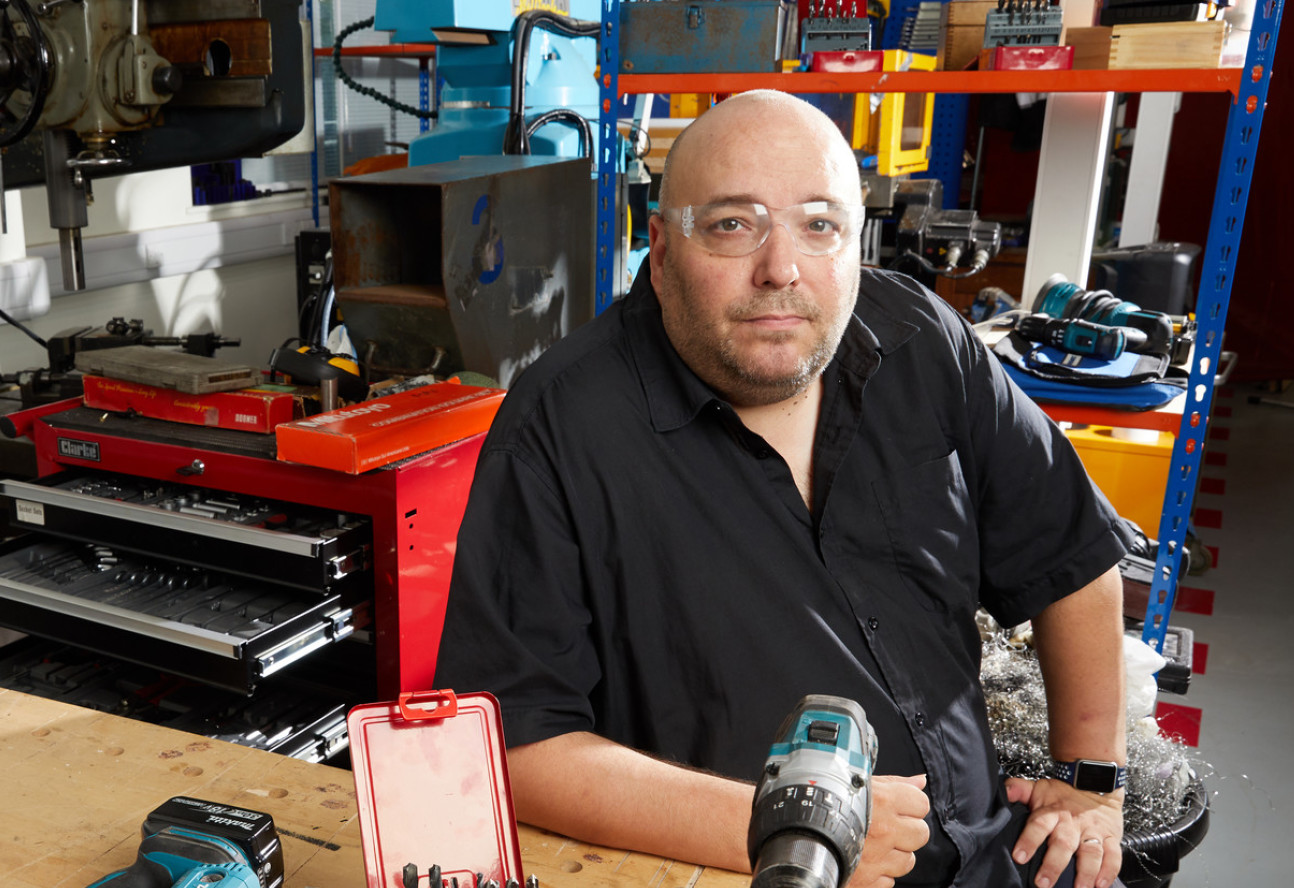 Bald man in black shirt and safety goggles sitting at bench in workshop