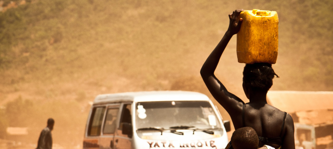 Photo of woman in Africa carrying water and walking towards a bus that is heading towards us the viewers.
