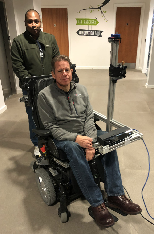 Photo of Team Captain Mahendran standing behind Paul in the gaze-led wheelchair. It looks like a regular wheelchair except there's a small bar in front of the face to track eye movements. 