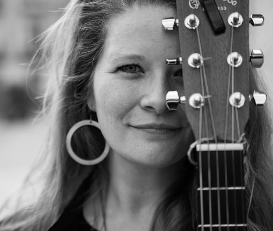 Black and white photo of Michelle Raymond, a woman with long hair wearing a hoop earring, with a guitar covering half of her face