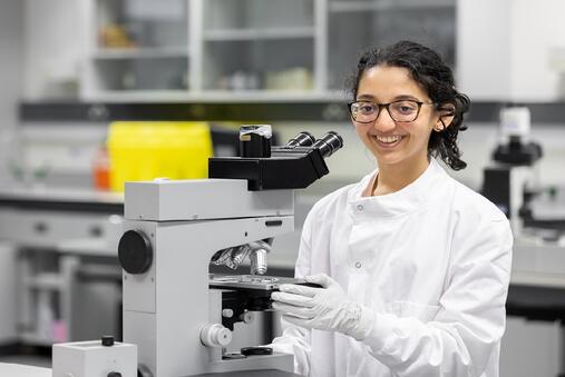 A young female student in a white lab coat with a microscope in front of her