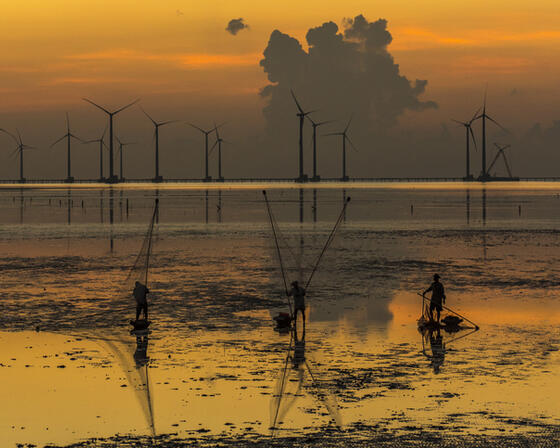 Sunset over the low tide at the Vietnamese seaside. Three fisherman sit in the foreground, with offshore wind turbines behind them in the distance