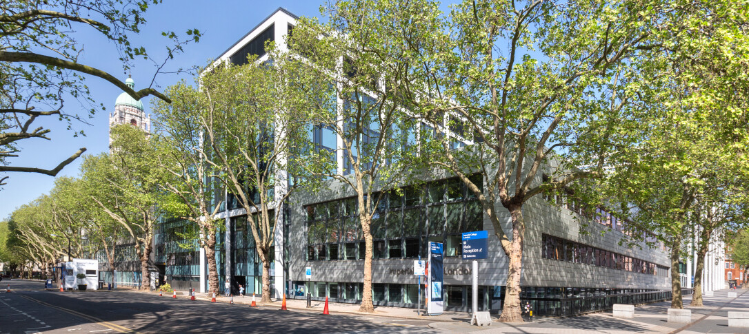 The exterior of the City and Guilds building from Imperial College Road on a sunny day. The trees on the road are blooming with green leaves, as if the photo was taken in spring. The sky is blue. The Queen's Tower is visible in the background. 