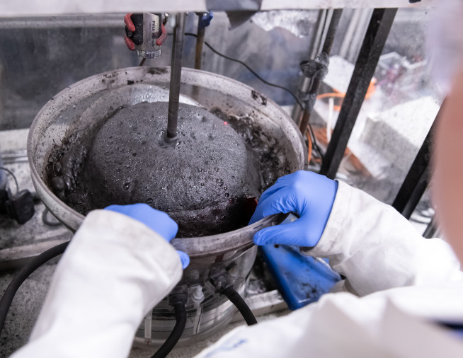 In a laboratory, a PhD student's gloved hands rest on the side of a bowl filled with grey slurry, part of a machine that separates minerals