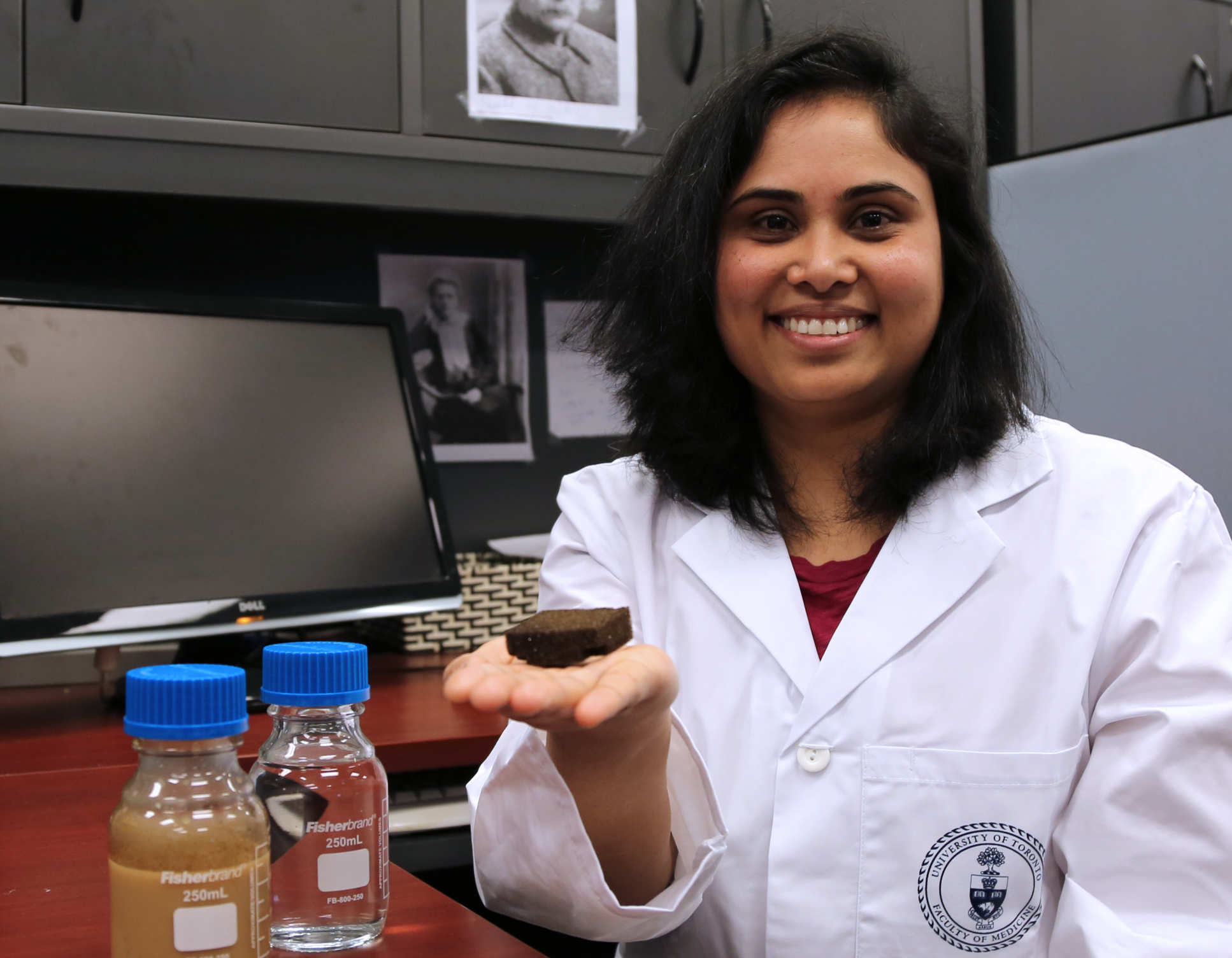Photo of Dr Cherukupally with the sponge and 'before and after' water samples (one bottle containing brown liquid; the other, clear, clean liquid after sponge treatment) in her lab at the University of Toronto