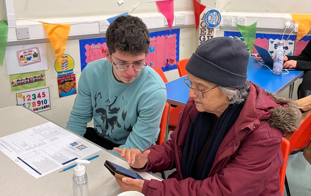A 'What the Tech?!' volunteer speaking to a participant who is using a smartphone.