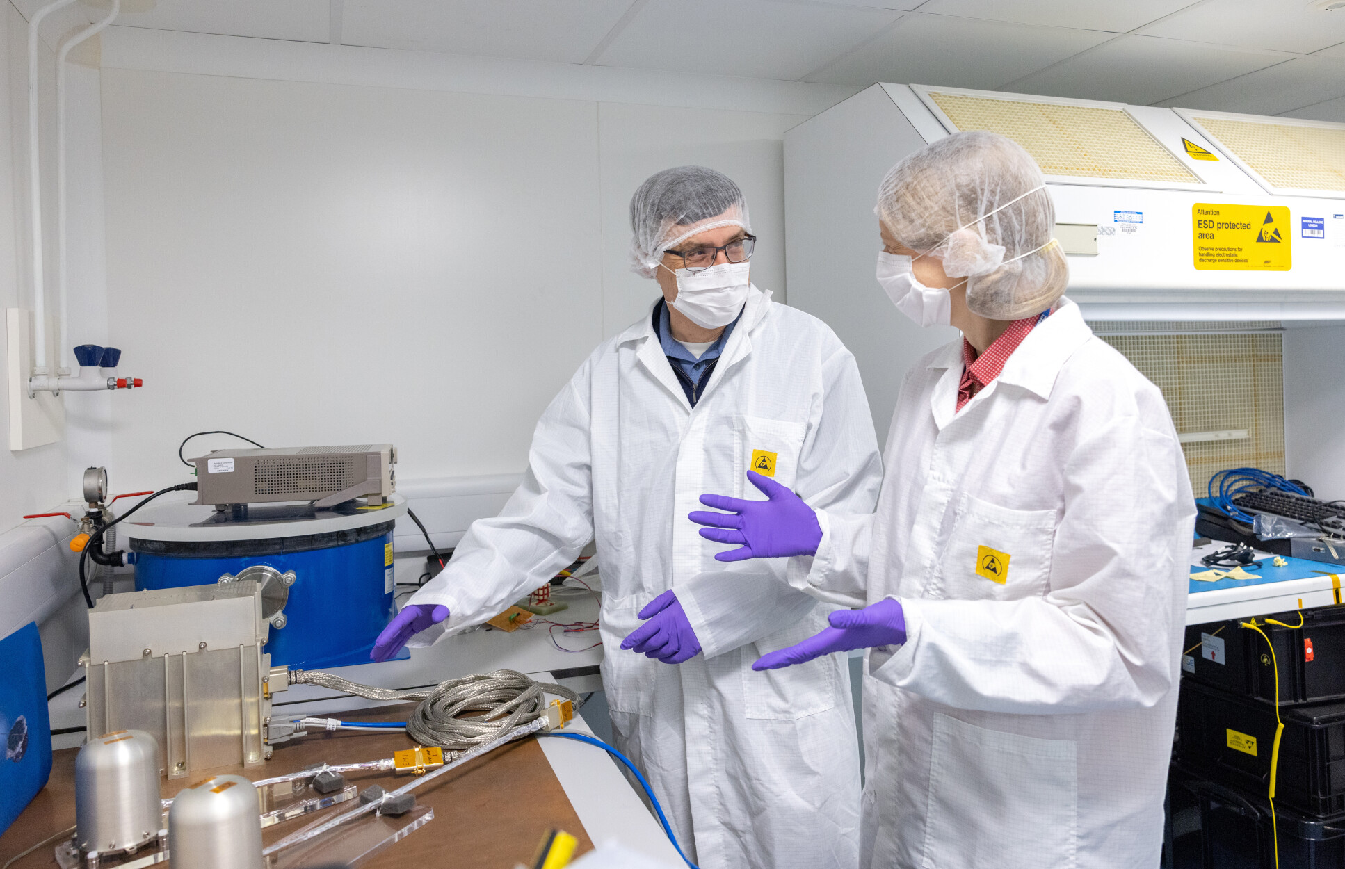 Two people in lab coats, gloves, masks and hirnets gesturing at equipment on a table