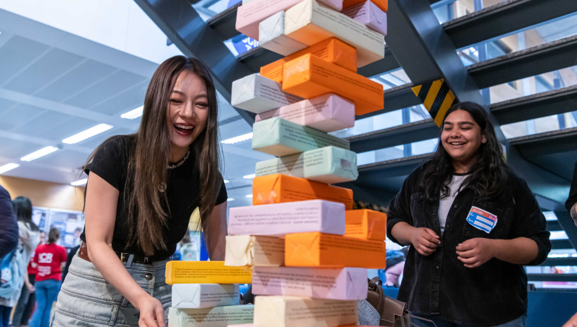 A girl with long brown hair laughing as she interacts with a giant jenga tower of pastel coloured bricks. Another girl looks on, smiling.