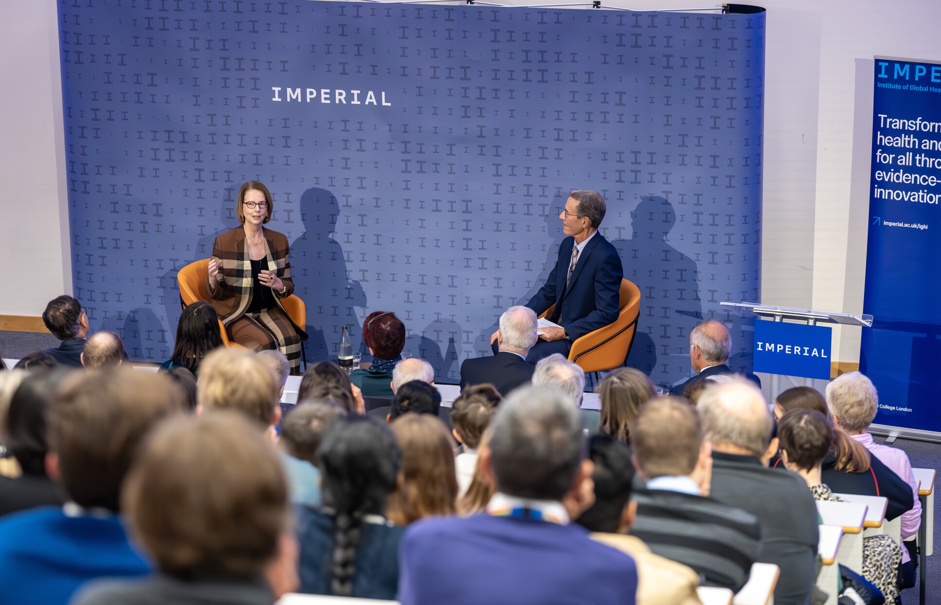 2 people sit on chairs speaking to a seated audience