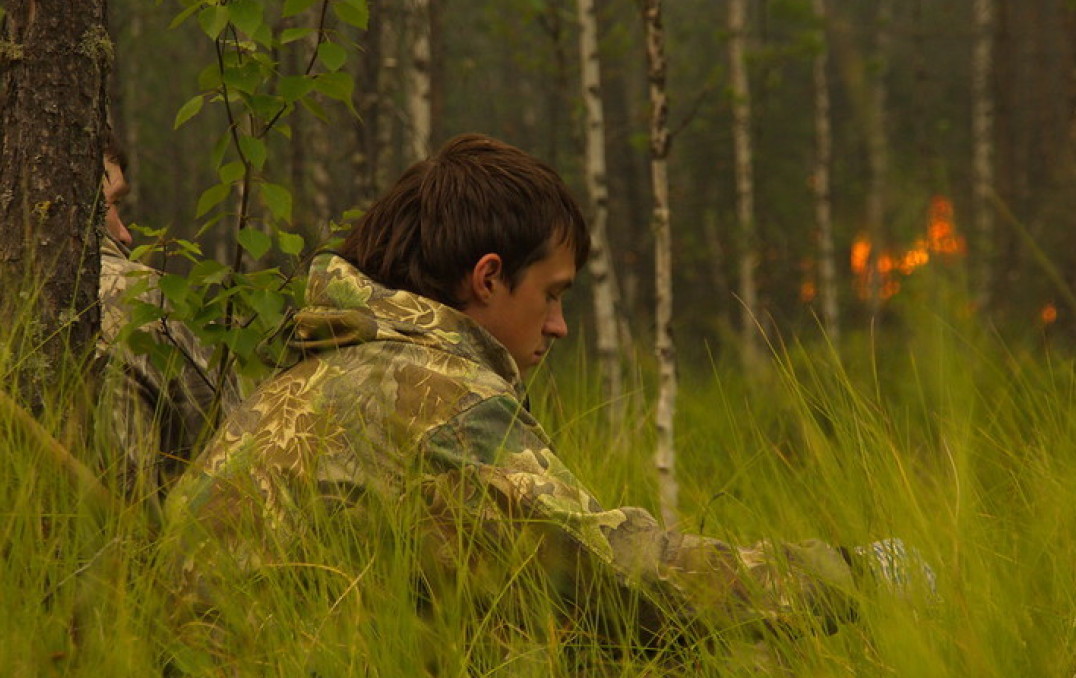 Young man sits in forest undergrowth as fire burns behind him