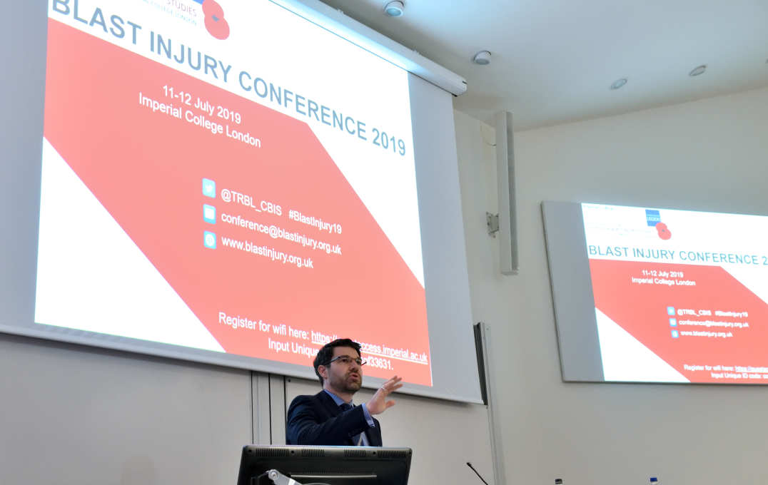 Dr Spyros Masouros, Chair of the Conference Committee, addresses the floor in front of two large screens with BLAST INJURY CONFERENCE 2019 on the slides