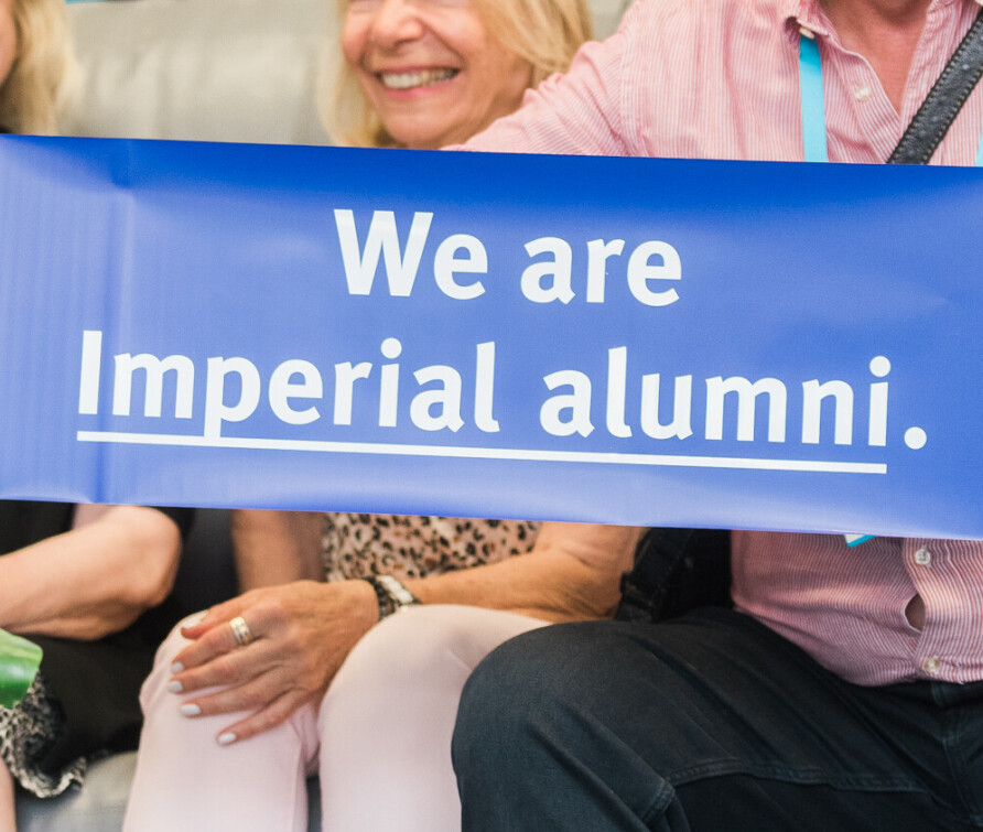 A group of three senior alumni, faces not visible, at an event. One holds up a purple pull-out banner that reads 'We are Imperial alumni.' The photo is cropped so that only their hands and part of their upper halves are visible. 