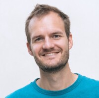 Headshot of Balazs Csuvar smiling. He is against a light grey background and wears a teal t-shirt.