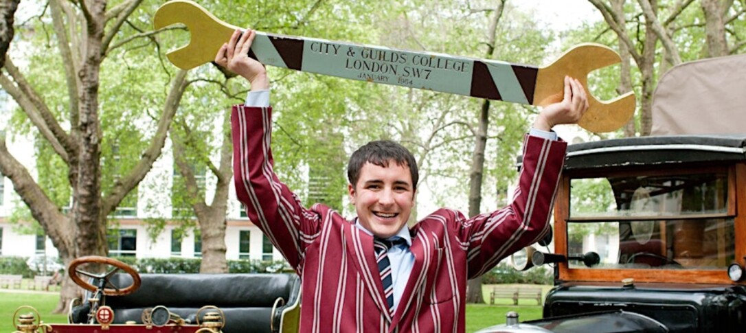 A young CGCA member wearing a burgundy striped uniform holds up a large city and guilds spanner prop. Boanerges the mascot is pictured in the background. 