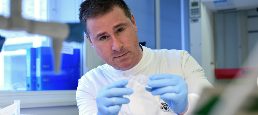 Portrait of Prof Darryl Overby in a laboratory. He is sat down and looking into the camera. He is wearing full PPE and holding a sample. 