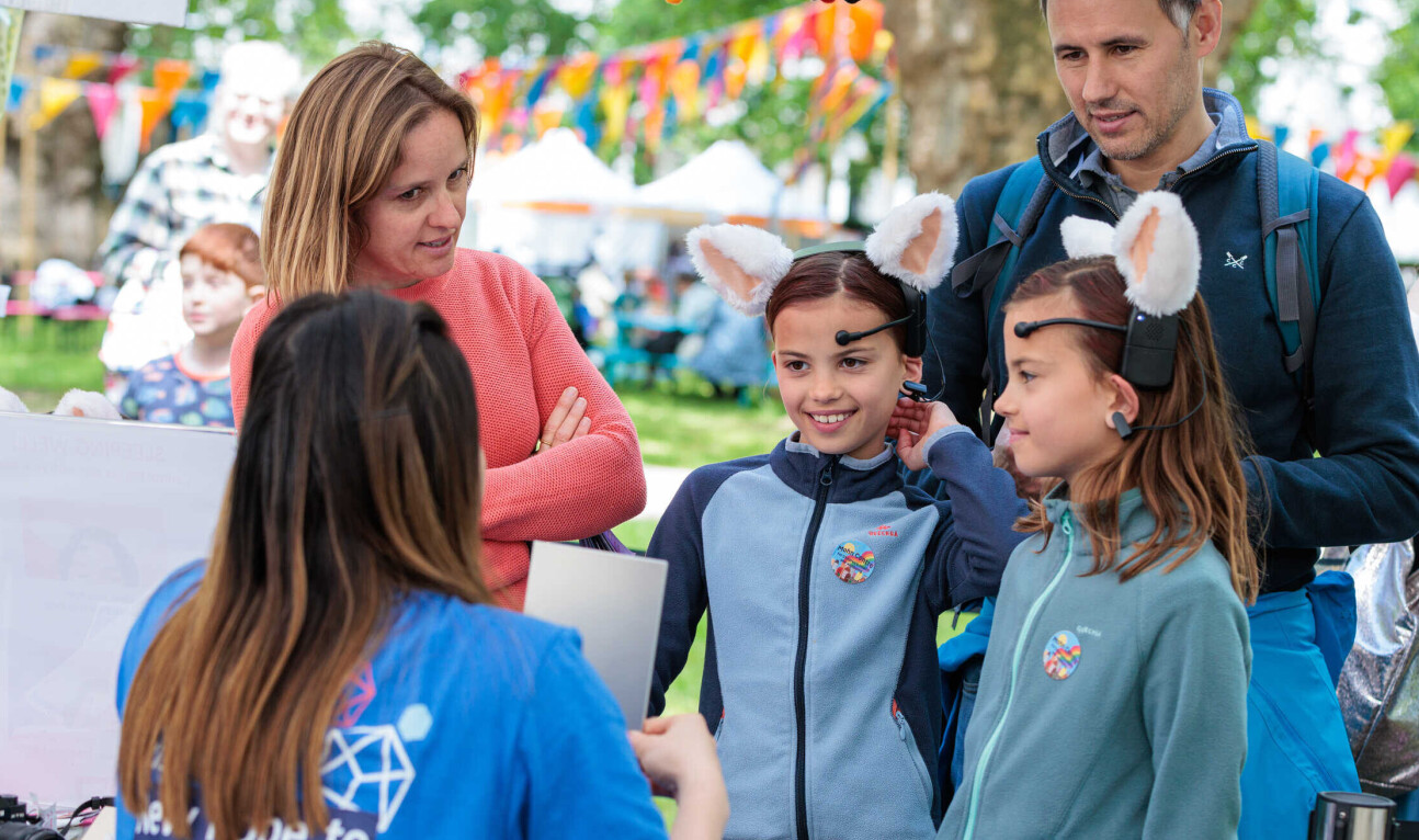 Two young girls, both smiling and wearing headbands with large, fluffy animal ears, stand beside their parents. A person in a blue t-shirt, facing away from the camera, is engaging with the family from behind a table.