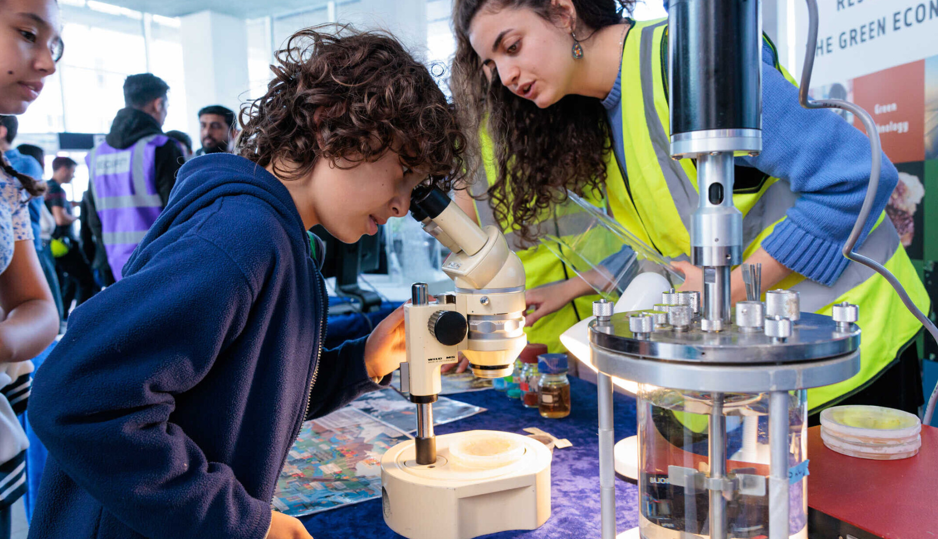 A young boy looking through a microscope at a science booth while a woman in a high-visibility vest, possibly a scientist or facilitator, helps him. The woman is leaning in, explaining something, while another girl stands nearby, watching attentively.