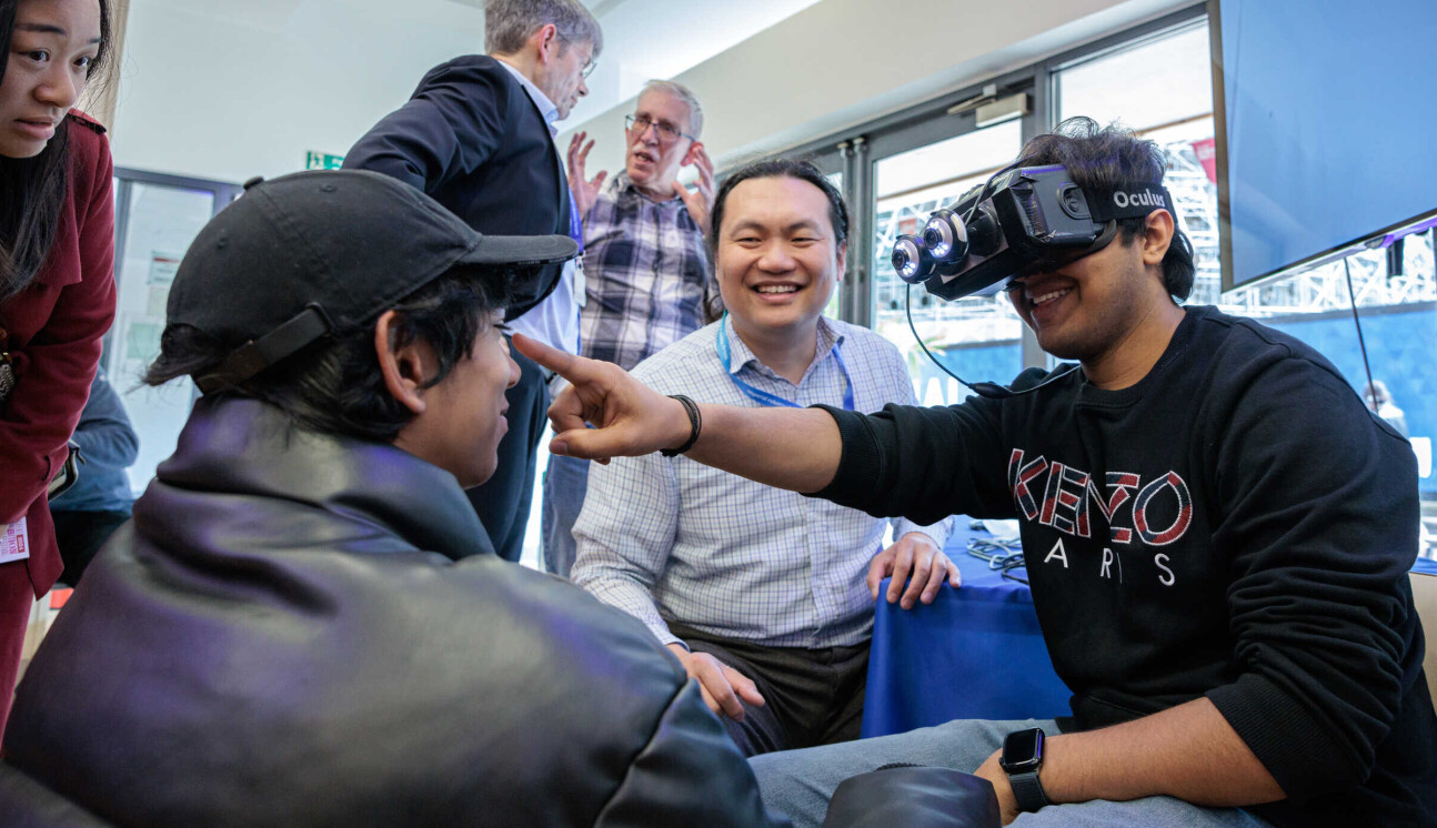 A man is smiling and wearing a VR headset. He is pointing ahead of himself, while two other men look on smiling.