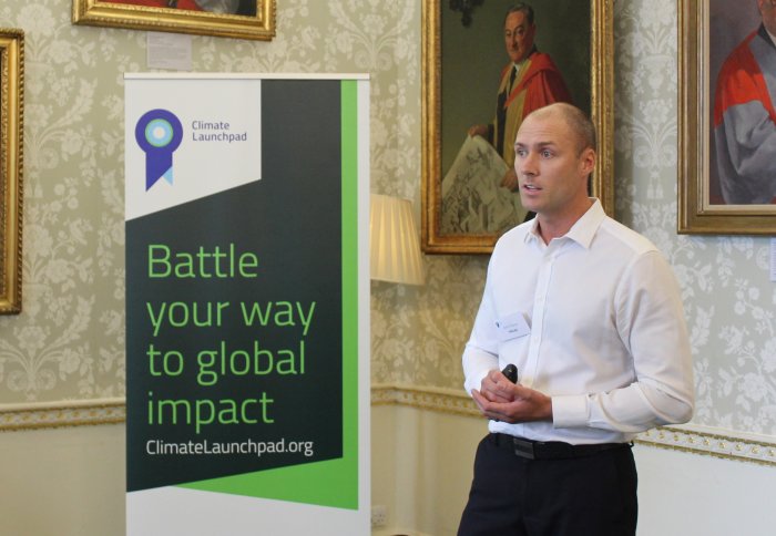 A man in a white shirt presents in front of a sign reading "Battle your way to global impact"