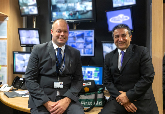 Two men in suits with blurred CCTV screens in background smile together