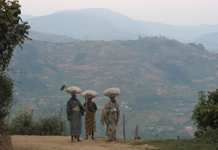 Three Ugandan women walk with bags on their heads, a mountain in the background