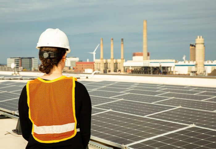 Worker installing solar panels with a power plant and wind turbine in the background