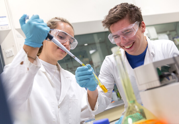 Two undergraduate students in lab coats and safety goggles, in a lab, pipetting a coloured solution into a test tube.