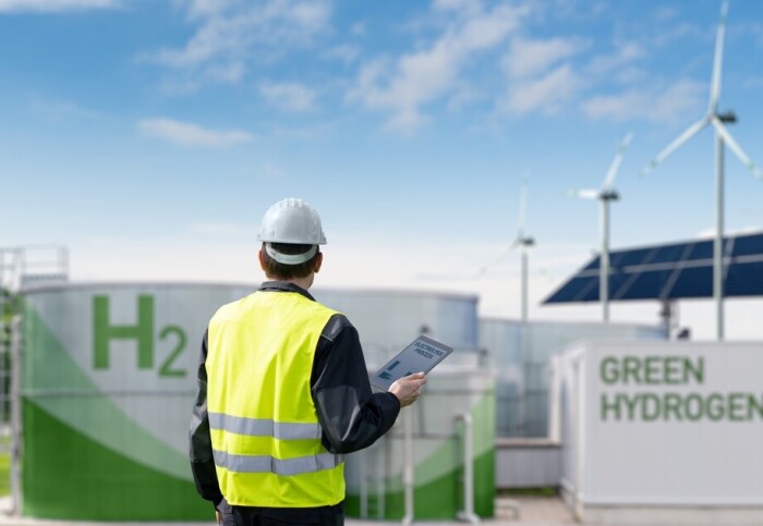 A contractor stands before two wind turbines and an energy storage system labeled 'Green Hydrogen'.