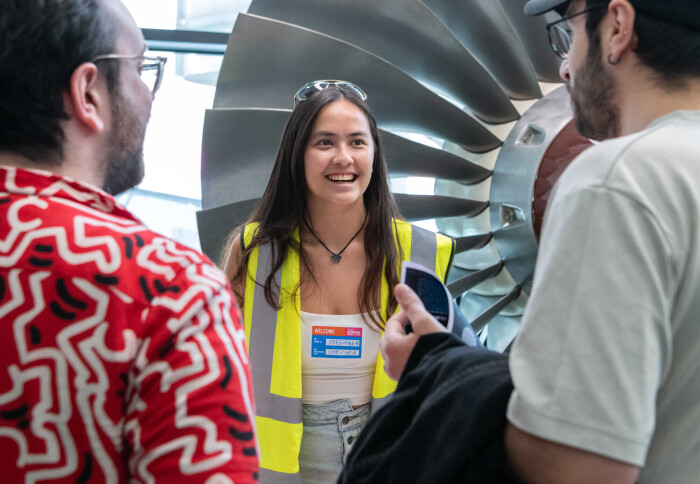 This image shows a young woman in a high-visibility yellow vest smiling and speaking with two men. They are all in front of a large industrial-looking turbine.