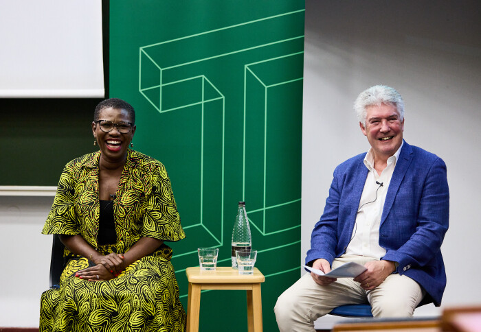 Two people sitting in front of a green banner with a large letter I on it smile