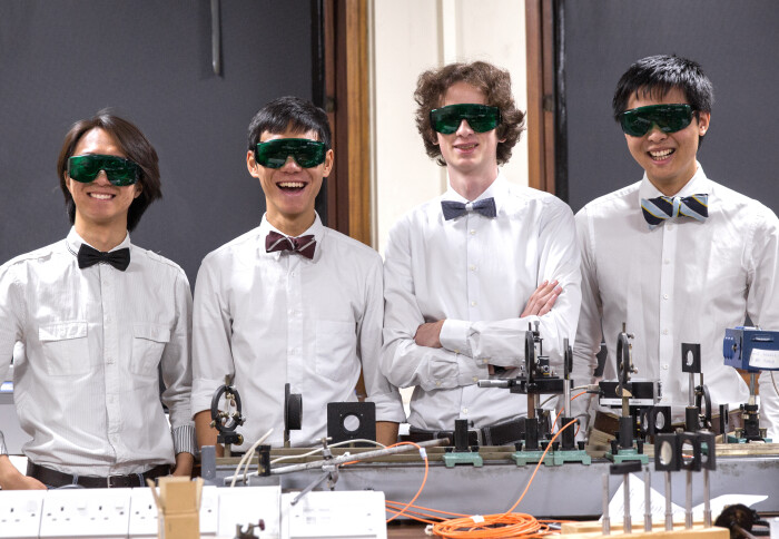 four engineering students smiling, wearing tinted lab googles and stood behind a table with equipment on