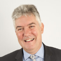 Headshot of Professor Frank Kelly, cheerfully smiling at the camera against a plain white background. He wears a light blue shirt and patterned tie with a charcoal coloured suit jacket.