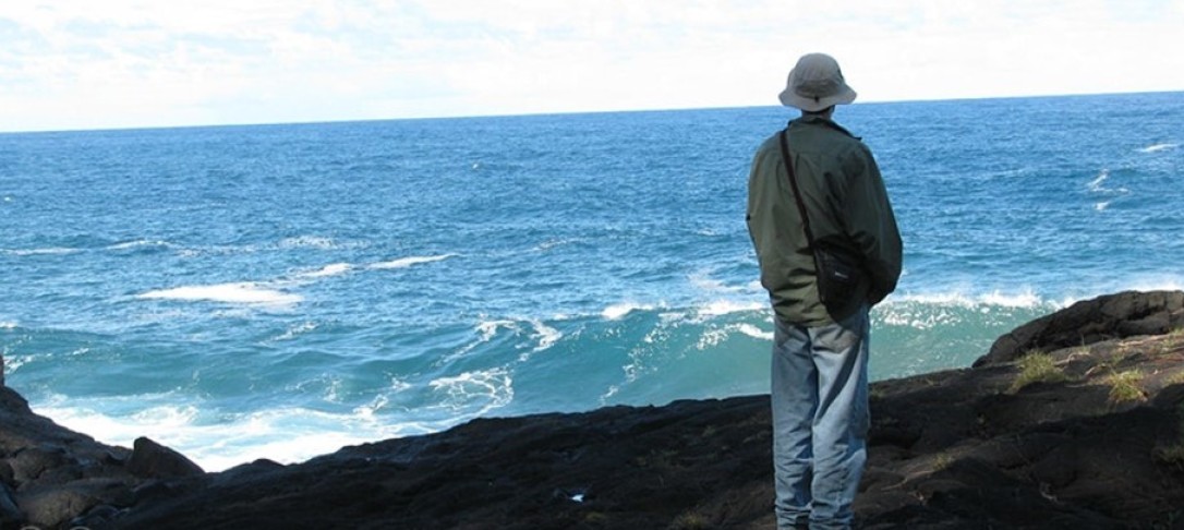 Man in hat giving us his back and standing on a cliff staring at the ocean and crashing waves against the rocks below