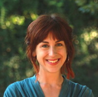 Headshot of Dr Sarah Holliday, she is pictured in front of a green background that looks like trees. The background has a high bokeh. Sarah is smiling and wearing a teal v-neck outfit.