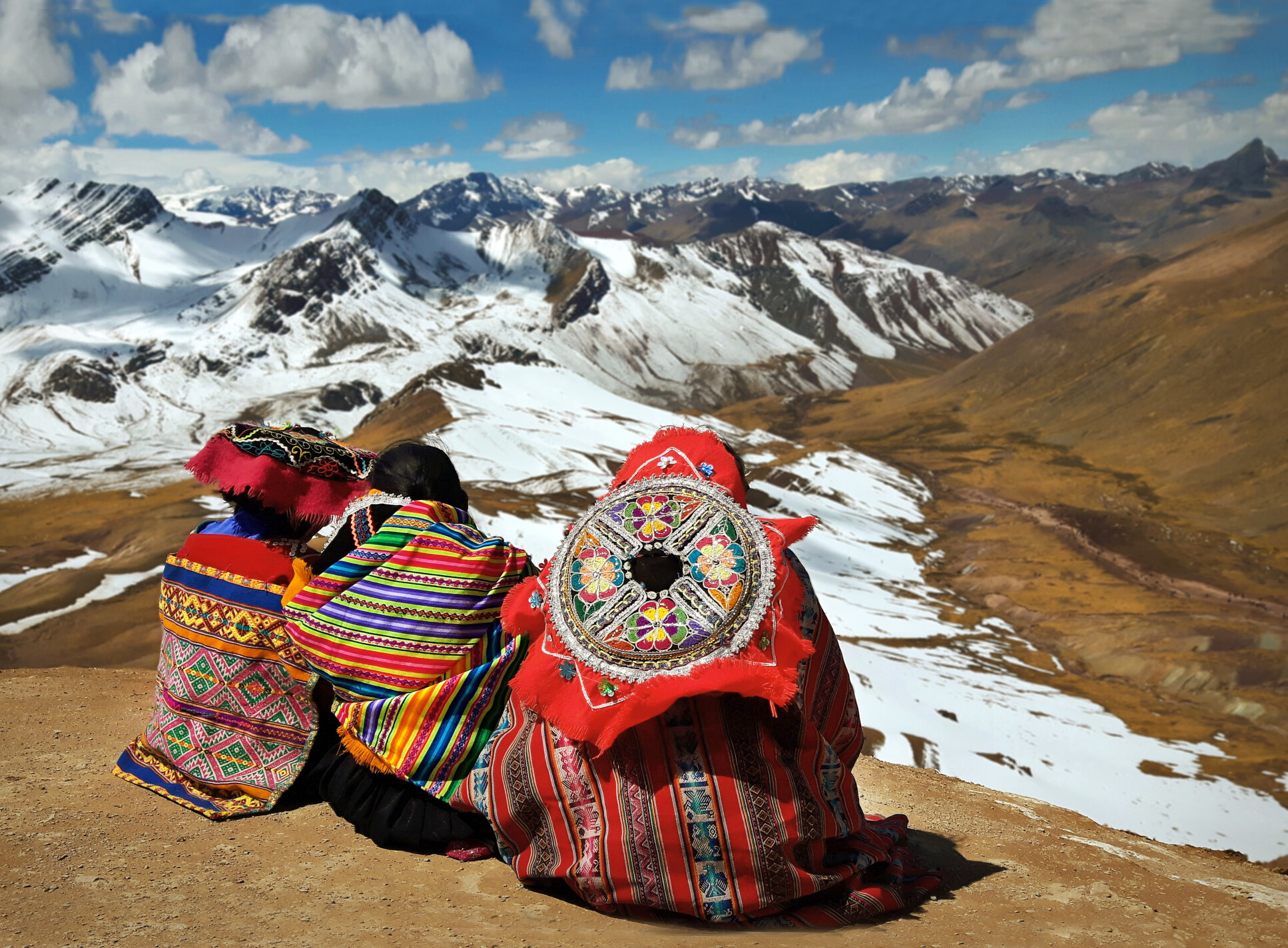 People contemplating the Andes in Peru. They sit on a peak, and look out over the snowy mountainscape.