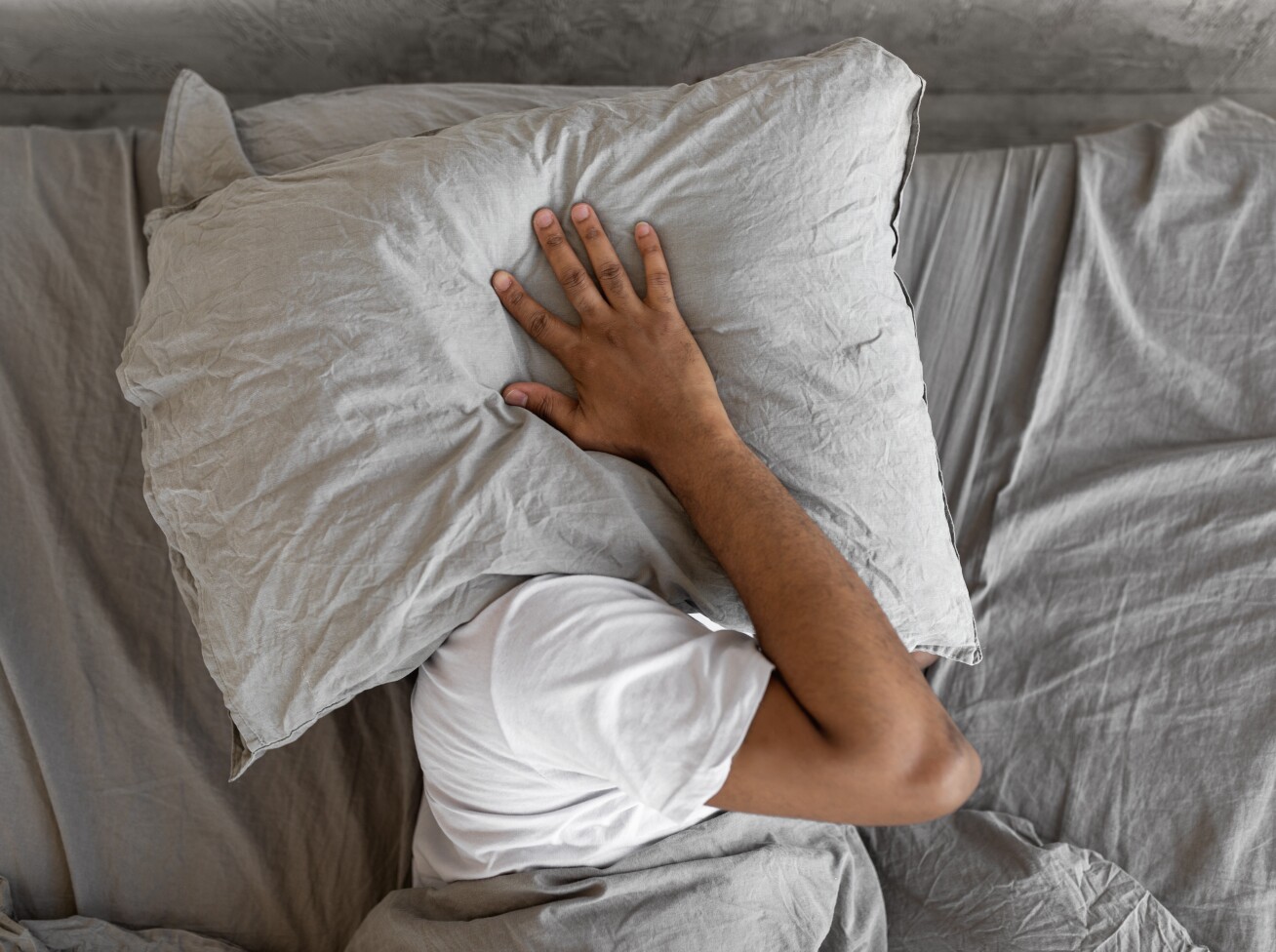 A man has a pillow over his head in bed because it is too loud