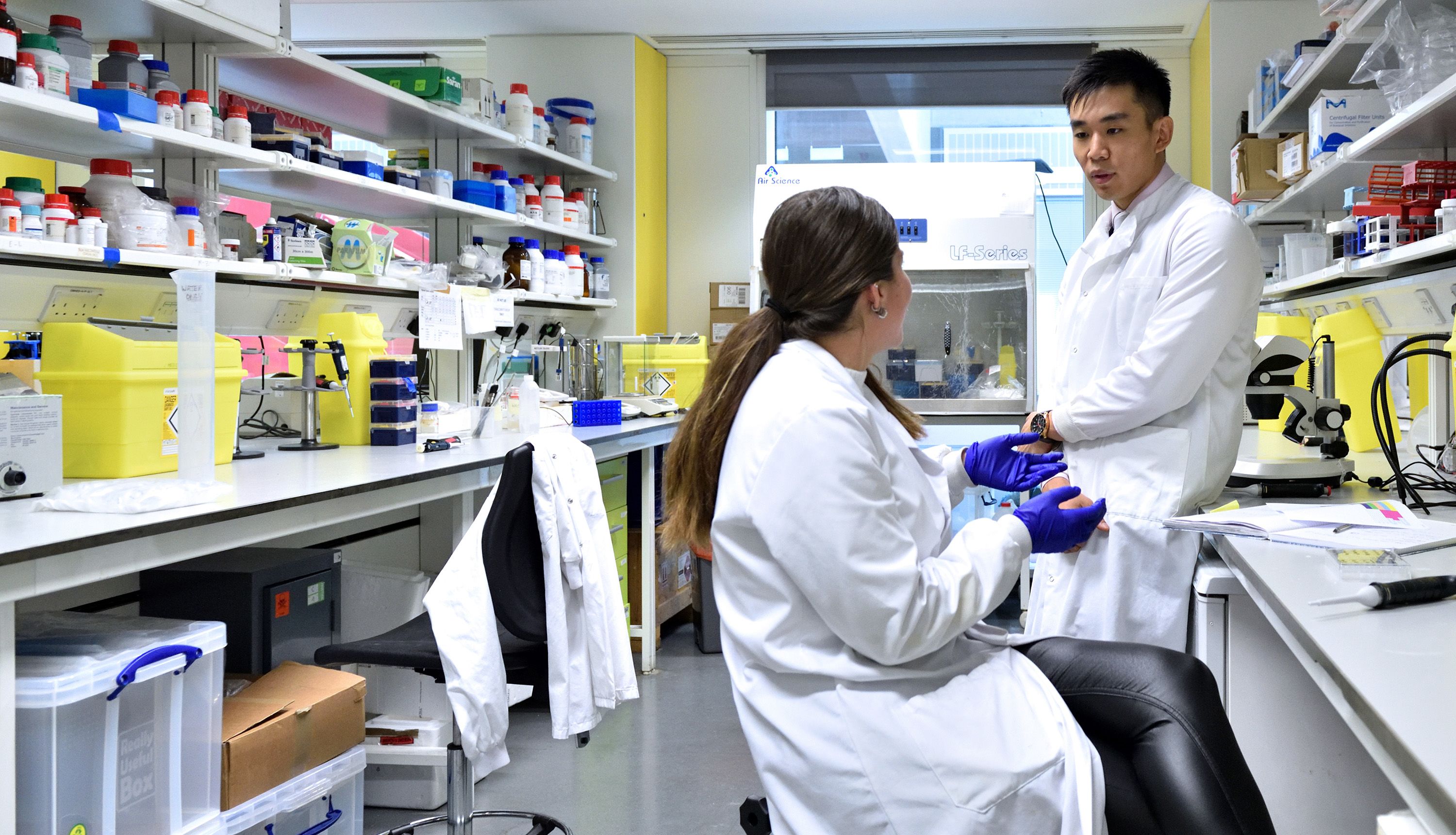 Brian wears a white lab coat and leans against a work surface while in conversation with a female colleague