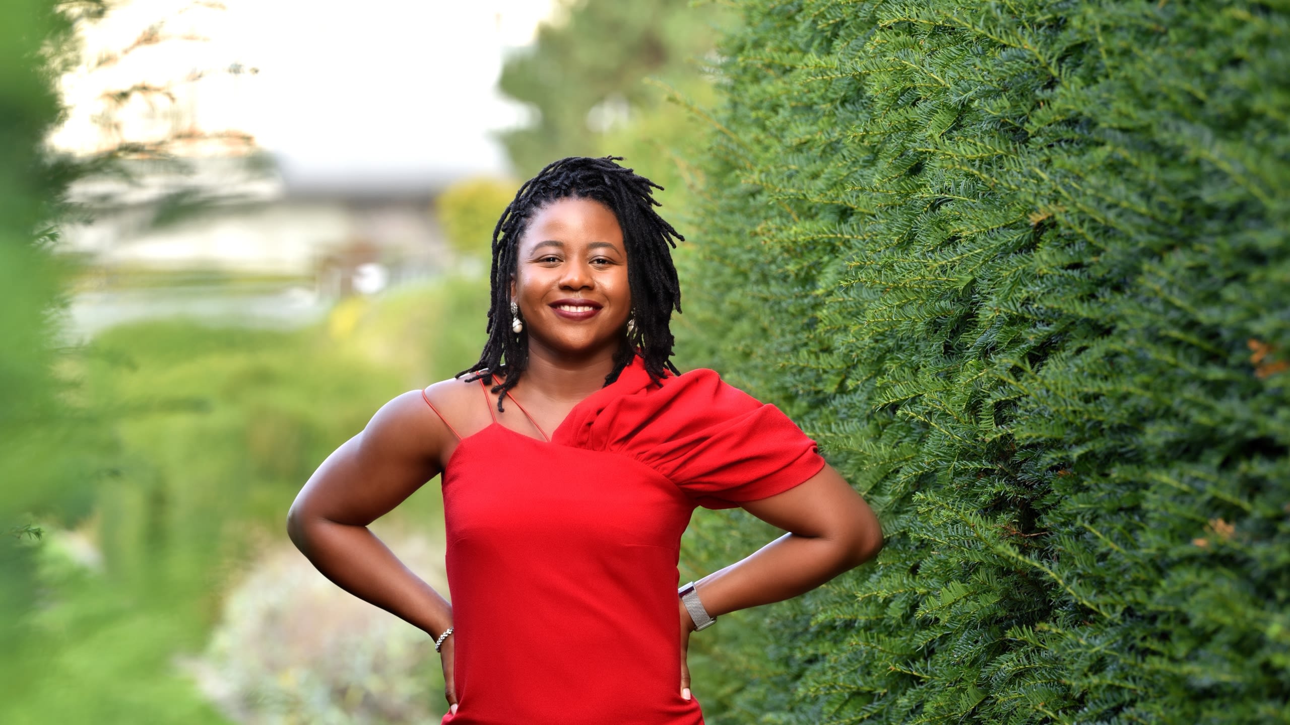 Dr Christiana Udoh standing by a large hedge with her hands on her hips, wearing an elegant red dress.