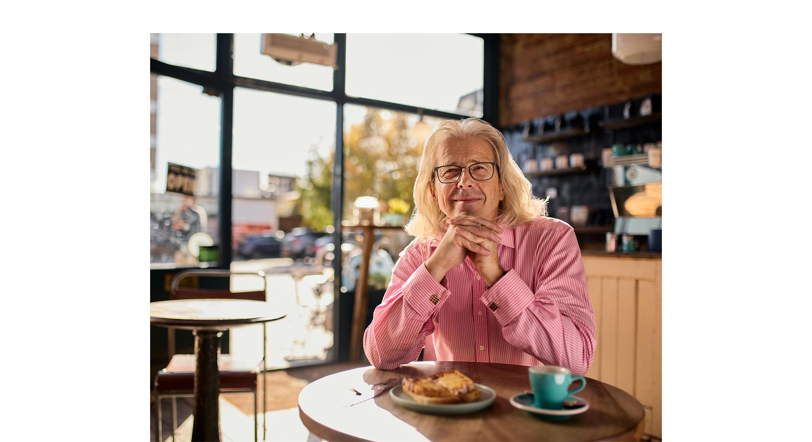 A blonde man wearing glasses and a red shirt sits in a cafe with a coffee and sandwich, looking to camera