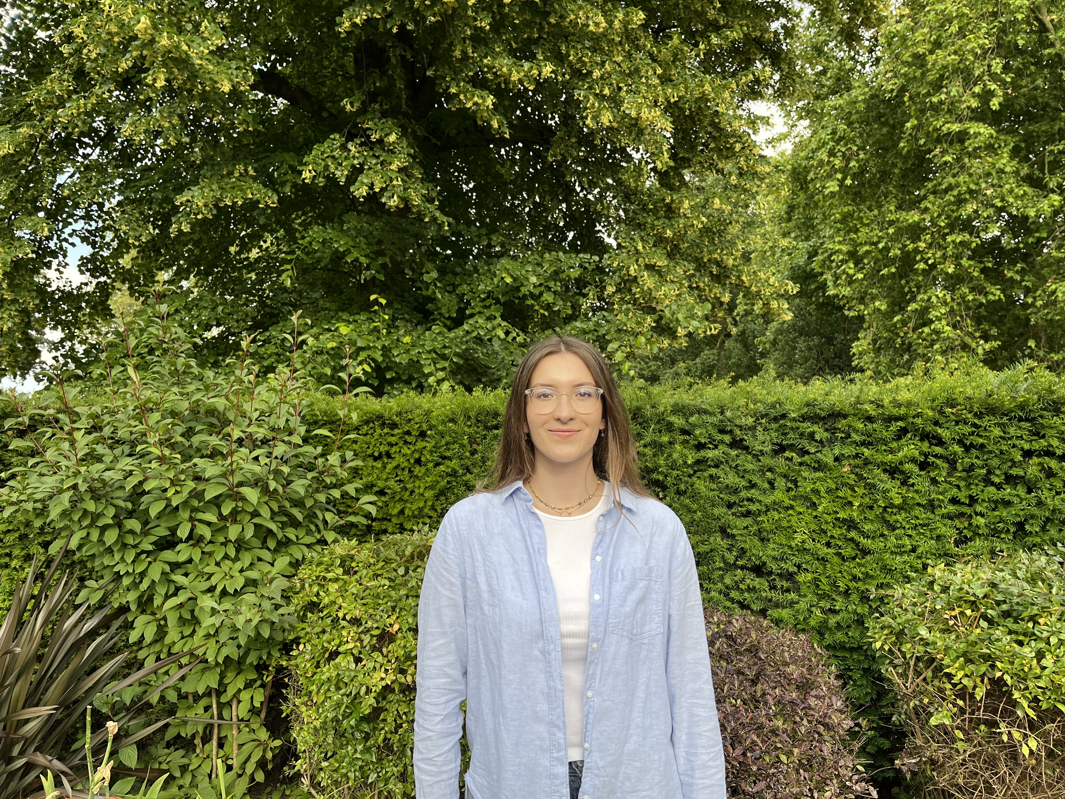 Profile photo of Alexandra wearing glasses and a white shirt with a light blue button down over the top, standing in front of a green hedge and trees