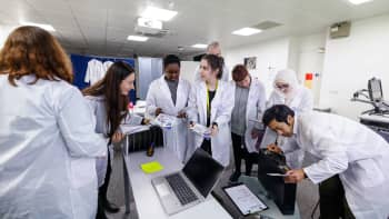 A photo of a group of people dressed in labcoats taking part in the PRESTIGE-AF escape room. The group are gathered around a laptop in discussion, whilst a team member is trying to solve the code to a locked briefcase.