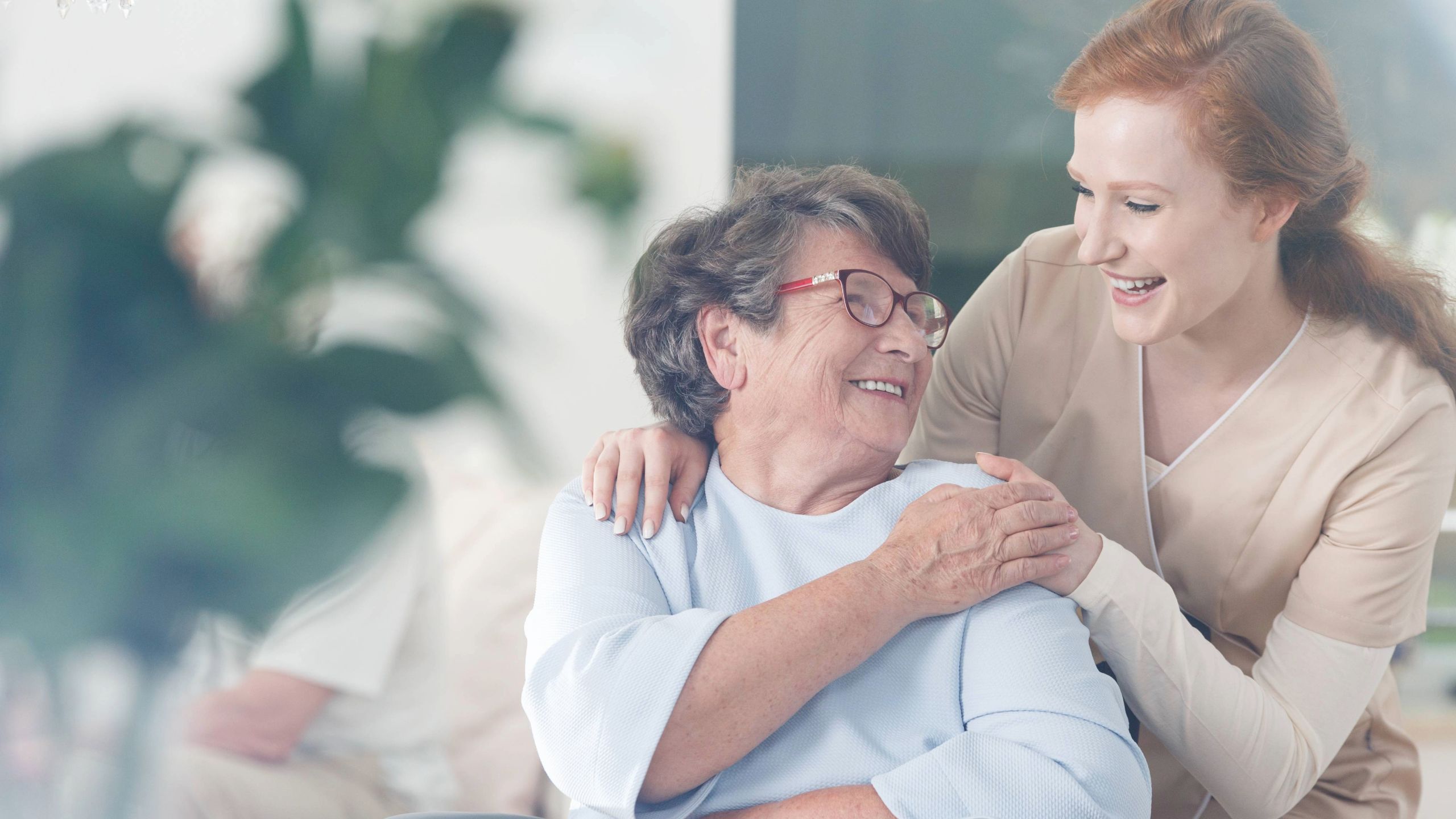 A photo of an elderly female patient and a female nurse looking at each other and smiling whilst holding onto one another in a supportive manner.