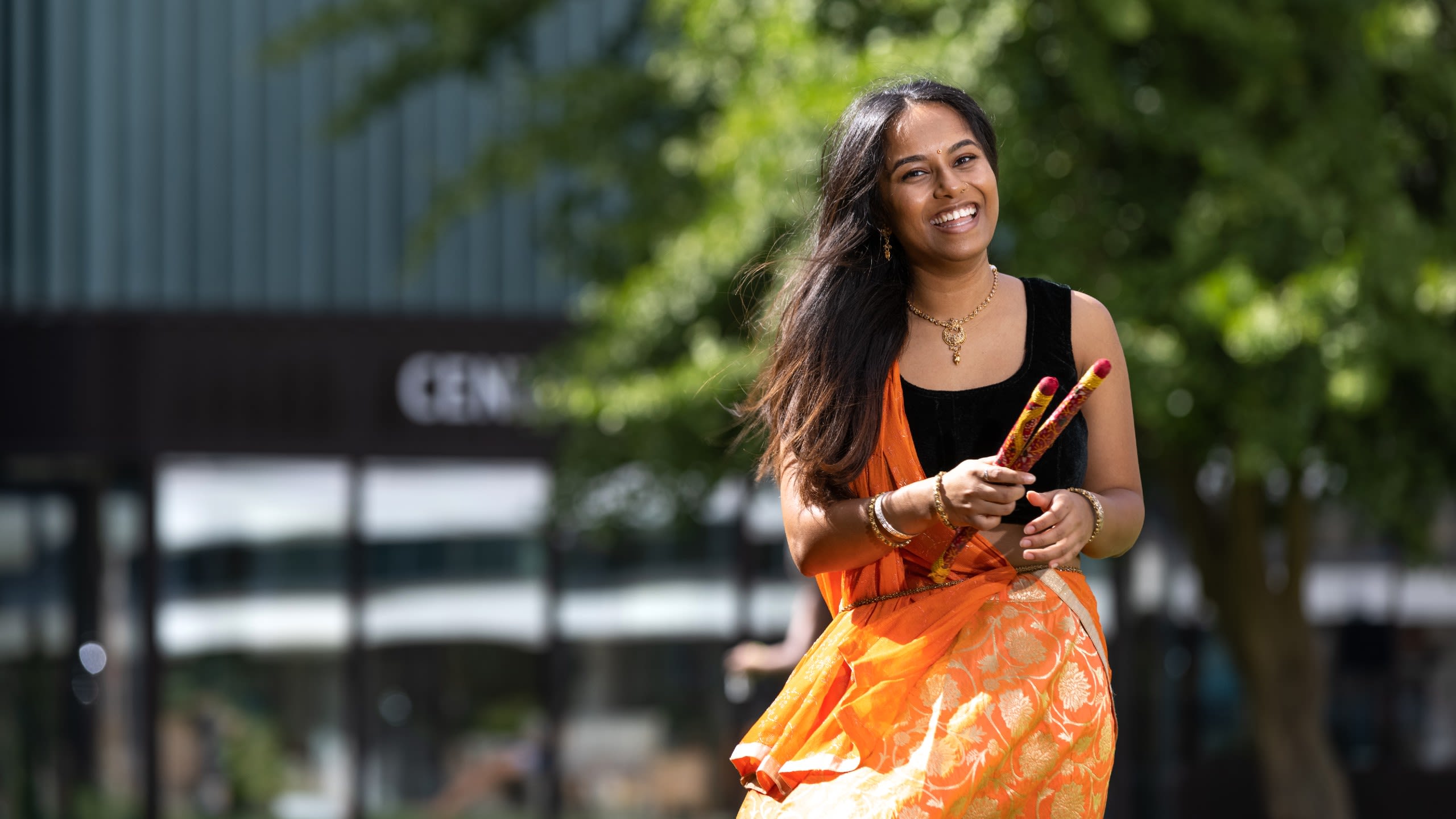Nandini Bhudia wears a South Asian 'lengha' outfit (black crop top and orange embroidered skirt with a long scarf) and holds two dancing sticks used to perform a cultural dance