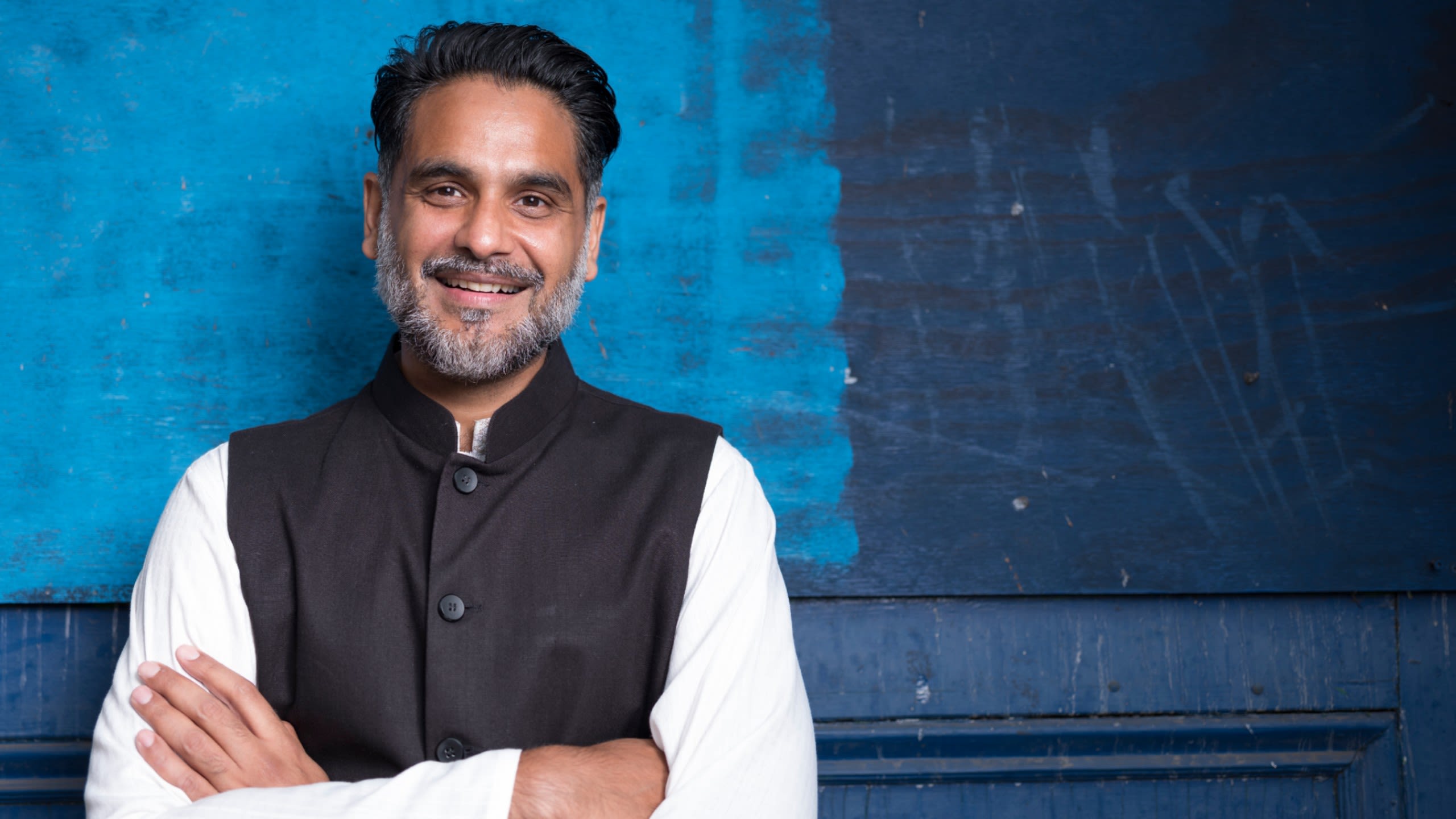 Syed pictured in traditional South Asian ethnic wear called 'shalwar kurta.' He stands against a vibrant blue wall with arms folded and smiles at the camera.