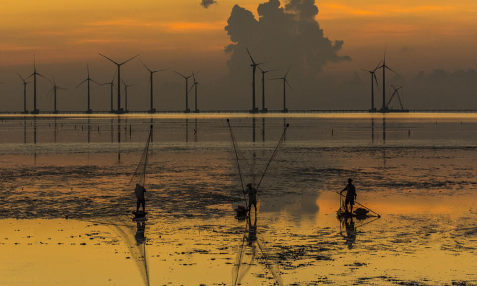 Sunset over the low tide at the Vietnamese seaside. Three fisherman sit in the foreground, with offshore wind turbines behind them in the distance