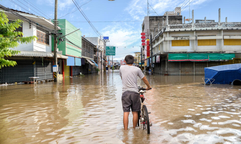 A man stands knee-deep in brown flood water covering an intersection of roads in Thailand.Shop fronts are closed to the public, powerlines hang in the distance