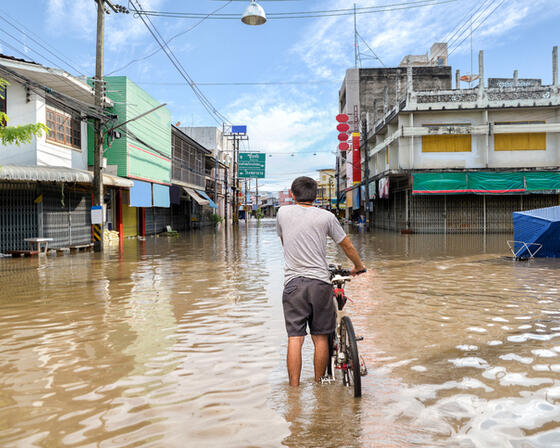 A man stands knee-deep in brown flood water covering an intersection of roads in Thailand.Shop fronts are closed to the public, powerlines hang in the distance