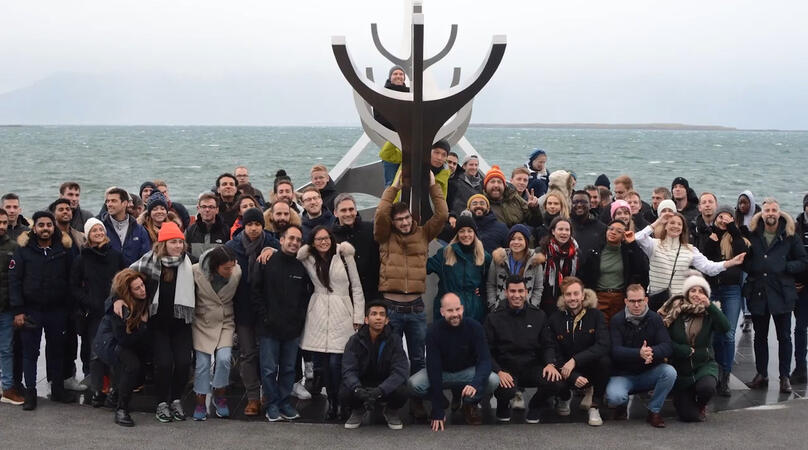 The WMBA cohort gathered together in front of a beach front in Iceland with an anchor in the middle