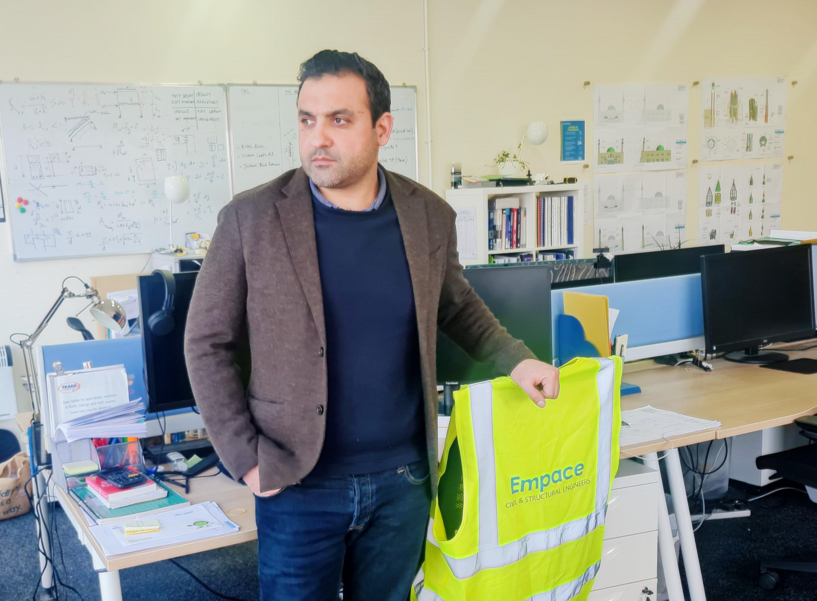 Ummer in his office at Empace, he is wearing a brown blazer over a black t shirt looking out towards where the window must be. He is learning on a chair which has a company high vis draped over the back