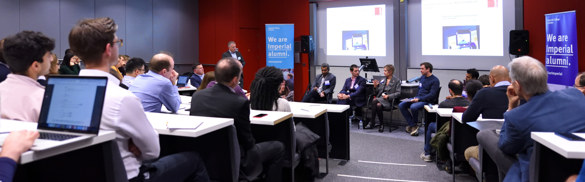 Busy lecture theatre focused on four speakers at the front of the room